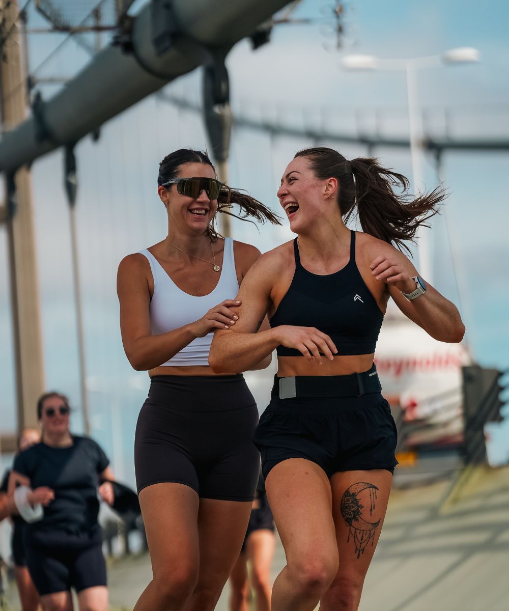 Two women laughing as they run along a sunlit path with other runners behind