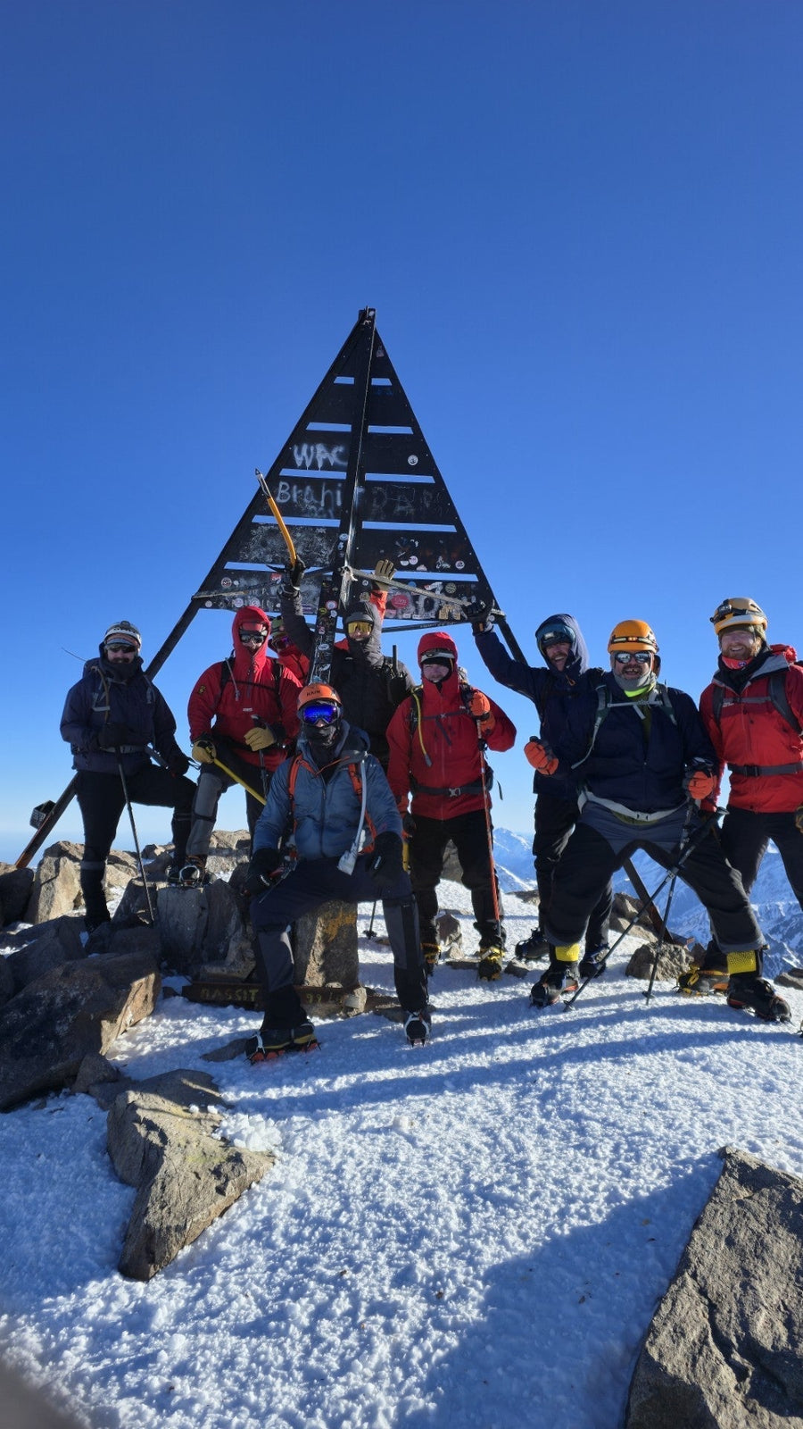 Group of climbers in winter gear at a snowy summit marker under blue sky